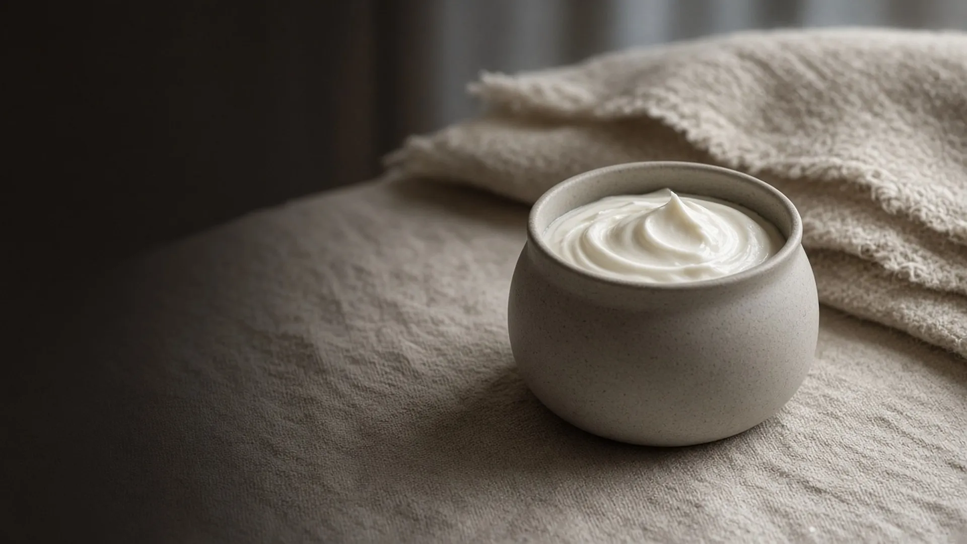 An open jar of rich cream on linen beside a wool throw